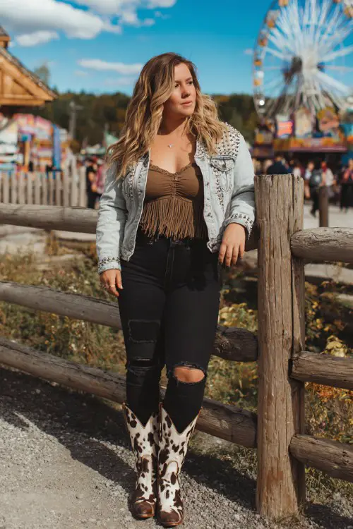A plus-size woman wears high-rise distressed black jeans with a tucked-in fringed suede crop top, layered with a studded denim jacket and cow print cowboy boots