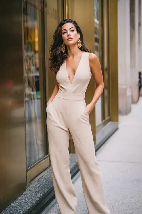 A tall, elegant woman wearing a beige ribbed sleeveless jumpsuit with a deep V neckline, fitted waist, and tailored straight-leg pants, hands in pockets
