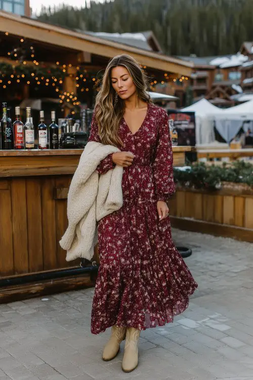 A woman in a wine-red long-sleeve floral maxi dress with a tiered skirt and subtle shimmer, styled with light beige cowboy boots and a soft ivory knitted shawl
