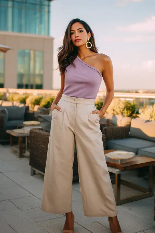 A woman over 30 wears a fitted lilac one-shoulder top tucked into high-waisted beige wide-leg trousers, styled with gold hoop earrings and tan block heels