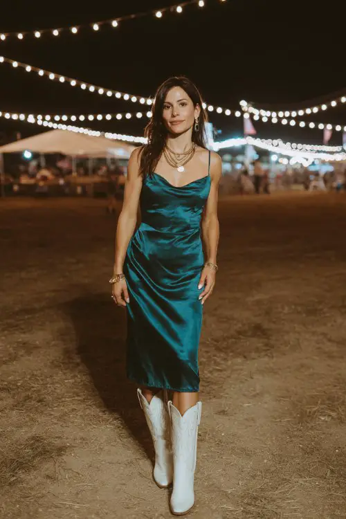 A woman over 30 wears a sleek satin midi dress in deep emerald, paired with white cowboy boots and minimal gold jewelry