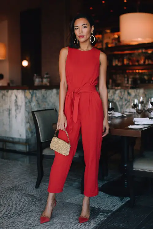 A woman over 30 wears a tailored red jumpsuit with a belt at the waist, gold hoops, and heels