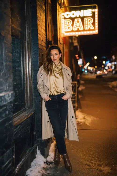 A woman over 30 wears dark skinny jeans with a silky blouse tucked under a long wool trench and heeled ankle boots