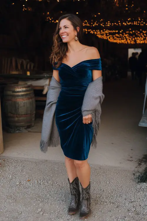 A woman over 40 in a dark teal velvet midi dress with a ruched bodice and off-the-shoulder neckline, styled with embellished cowboy boots and a gray cashmere shawl