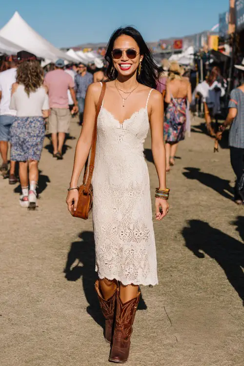 A woman over 40 wears a cream lace midi dress with spaghetti straps and scalloped hem, paired with brown Western boots, stacked bracelets, leather crossbody bag