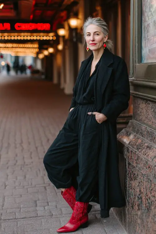 A woman over 40 wears a tailored black jumpsuit over red cowboy boots, layered with a long wool coat and statement earrings, posed outside a stylish theater venue