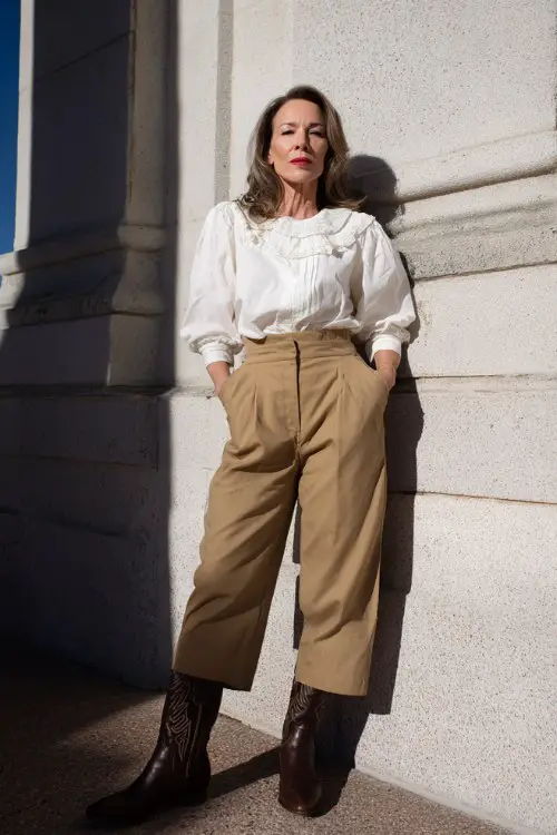 A woman over 40 wears high-waisted tan trousers with a white ruffle blouse tucked in and dark cowgirl boots