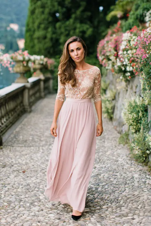A woman over 50 wears a flowy blush pink A-line wedding guest dress with lace bodice and sheer sleeves
