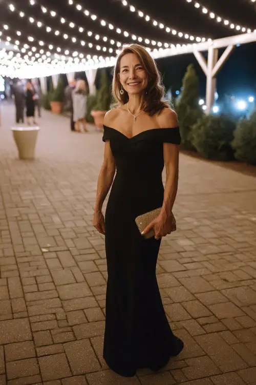A woman over 50 wears a long black off-the-shoulder evening gown with a fitted silhouette and small clutch