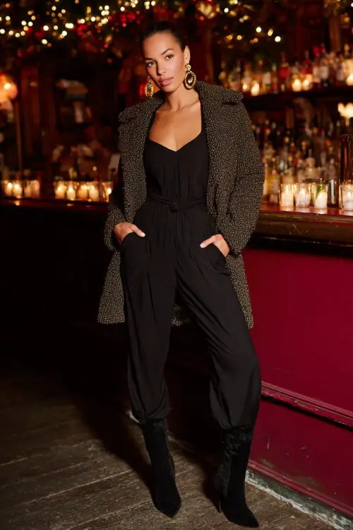 A woman wears a black jumpsuit with a belted waist and a teddy-textured overcoat, paired with bold gold hoops and heeled boots