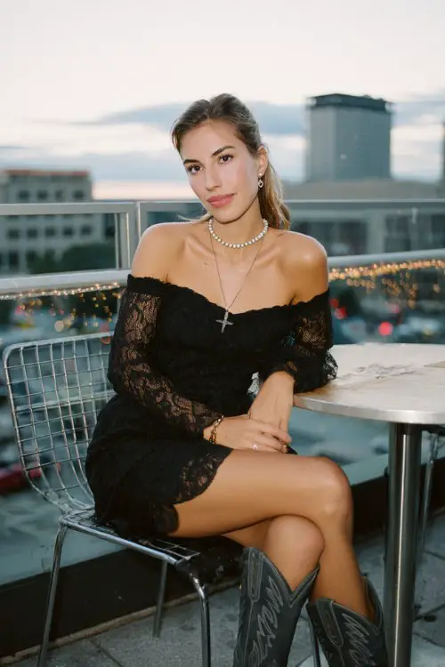 A woman wears a black off-shoulder lace dress with dark gray cowboy boots and a pearl choker