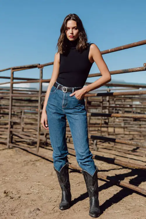 A woman wears a black sleeveless turtleneck bodysuit tucked into high-rise blue jeans with a statement silver buckle belt and black cowboy boots