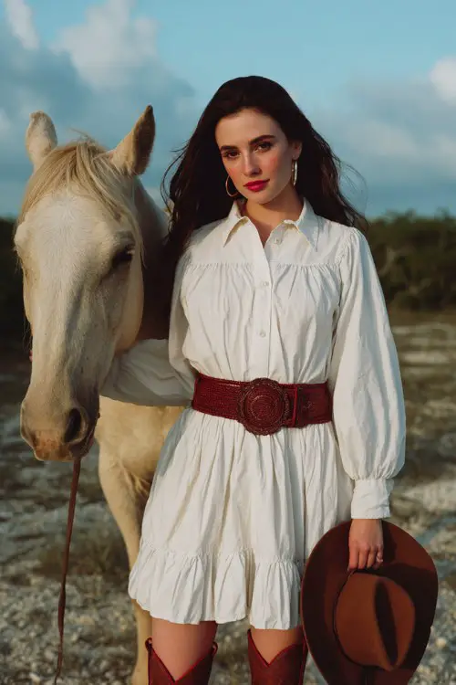 A woman wears a collared white shirt dress with ruffled hem and a wide brown woven belt, styled with red cowboy boots and silver hoop earrings, holding a cowboy hat