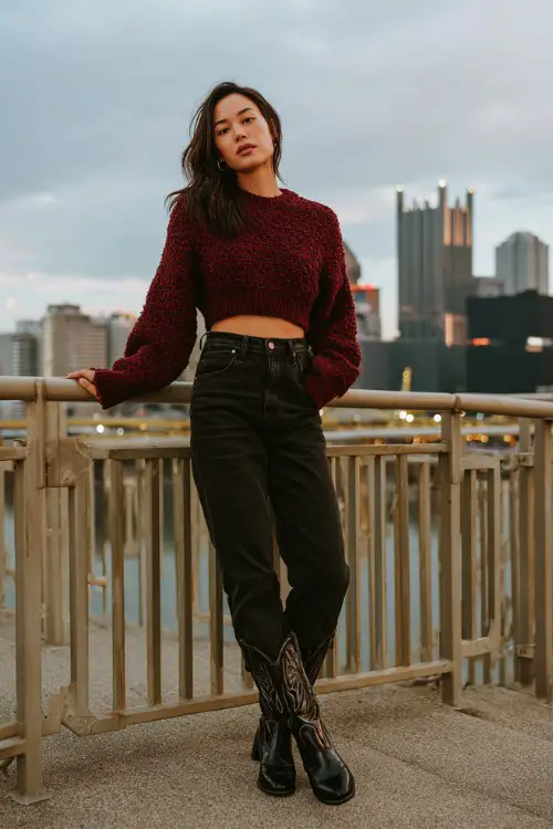 A woman wears a cropped burgundy sweater with high-waisted black jeans and glossy black cowboy boots