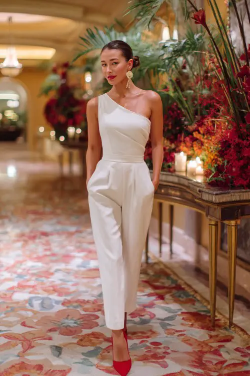 A woman wears a one-shoulder white jumpsuit with gold statement earrings and red heels