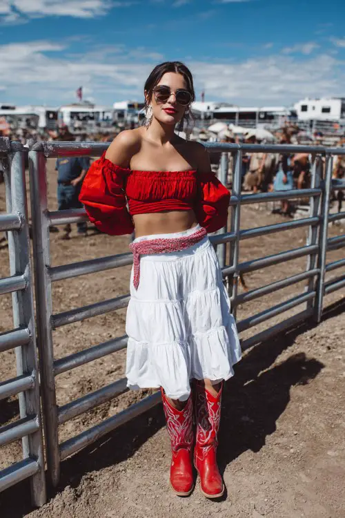 A woman wears a red crop top with puff sleeves, a tiered white skirt, and red cowgirl boots