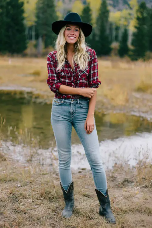 A woman wears a red plaid shirt with cuffed sleeves tucked into light blue skinny jeans, styled with short black cowboy boots and a black hat