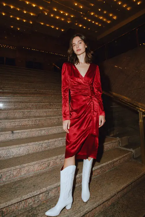 A woman wears a ruby-red satin midi dress with a ruched waist and pointed white cowboy boots