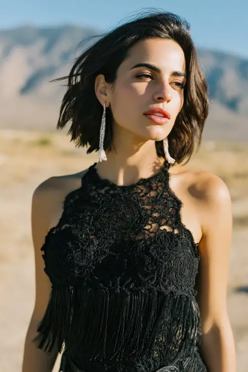 A woman wears a sleeveless black lace cocktail dress with dramatic fringe along the hem and black cowboy boots, accessorized with bold white tassel earrings