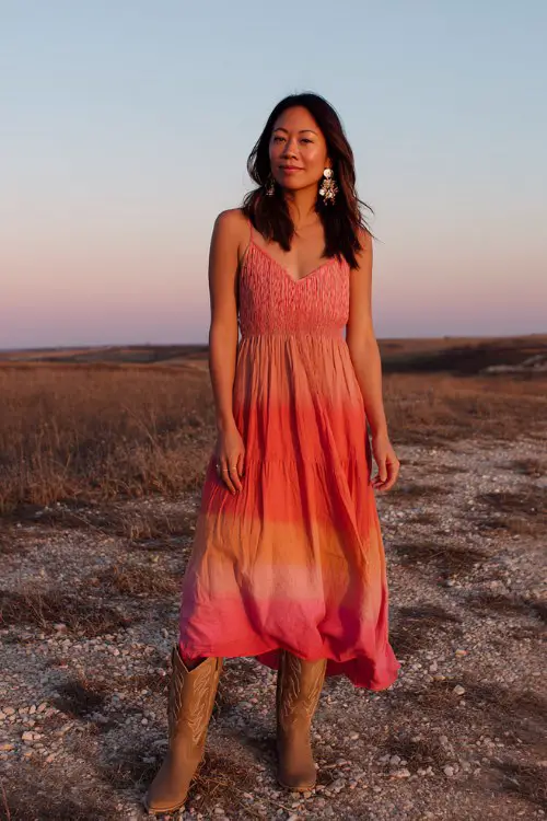 A woman wears a sunset ombré dress blending coral, pink, and orange tones, paired with light brown cowboy boots and bold earrings