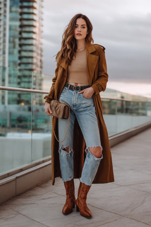 A woman wears caramel brown cowboy boots with ripped boyfriend jeans, a fitted crop tee and longline trench coat, styled with a chain belt and mini clutch