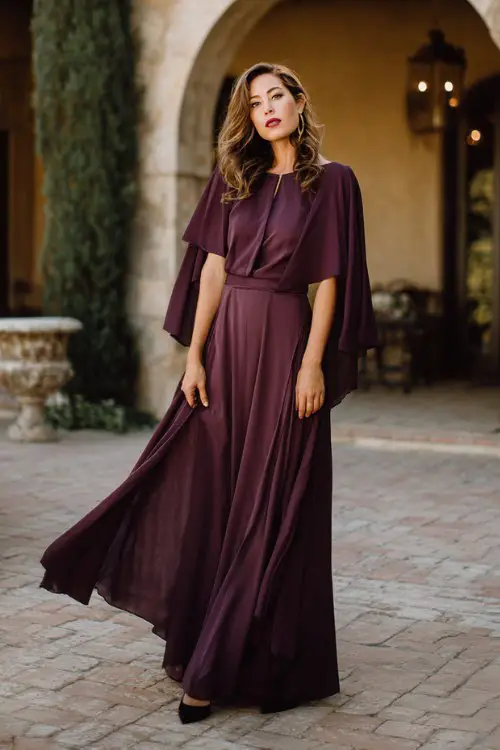 Timeless classy spring wedding guest fashion, woman over 40, plum purple flowing maxi dress with cape sleeves, black pumps
