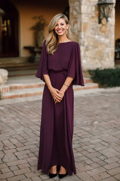Timeless classy spring wedding guest fashion, woman over 40, plum purple flowing maxi dress with cape sleeves, black pumps