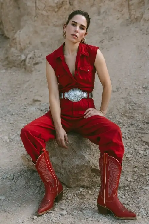 woman wearing a fitted red western jumpsuit with a deep neckline, oversized silver belt buckle, and bold red cowboy boots for a rodeo