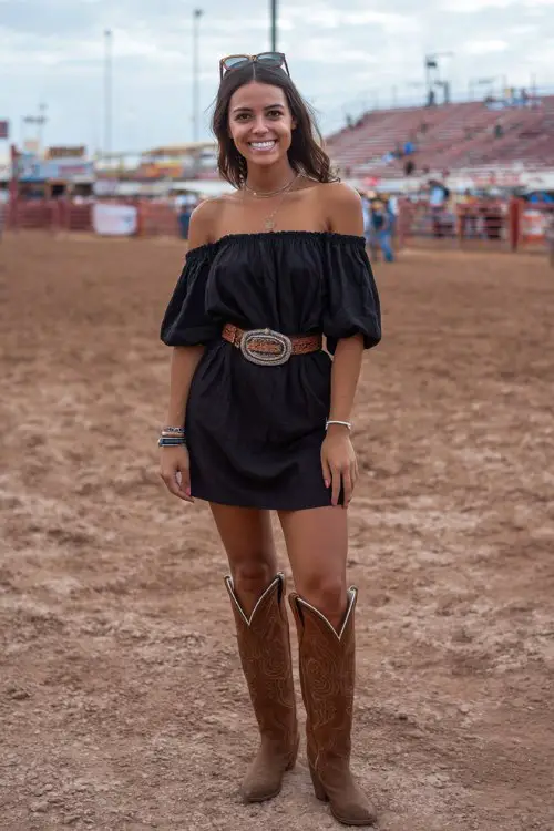 woman wears a black off-shoulder ruffle mini dress with a silver buckle belt and tall brown cowboy boots