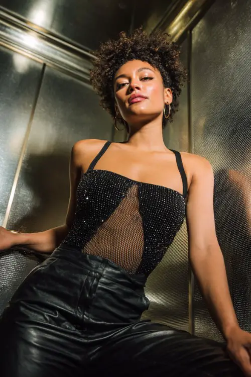 A Black woman with a short afro wears a black mesh bodysuit with rhinestone details, leather pants, and platform heels