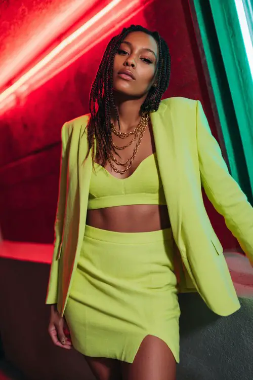 A Black woman with braids wears a lime green co-ord set featuring a cropped blazer and a mini skirt with side slits, paired with pointed pumps