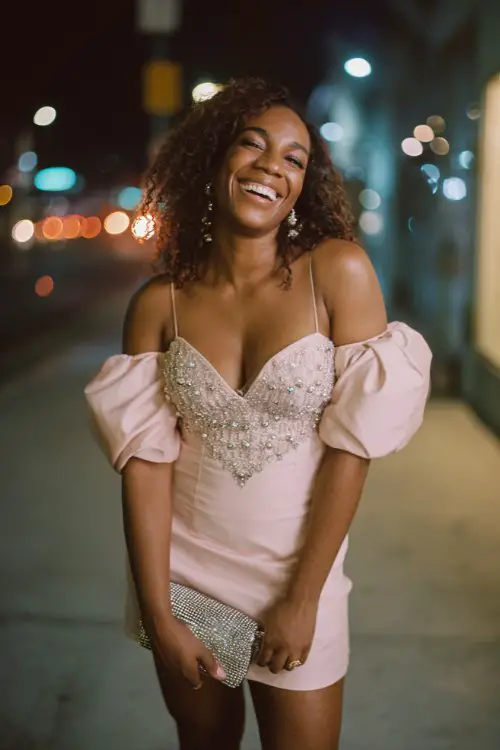 A curvy Black woman wears a baby pink structured mini dress with puffy sleeves and crystal embellishments, paired with clear heels and a sparkly micro bag