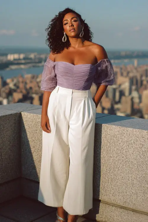 A curvy Black woman wears a pastel purple off-shoulder corset top and high-waisted white wide-leg trousers, styled with silver heels and hoop earrings