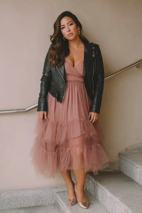 A curvy woman wears a dusty rose tulle cocktail dress with layered skirt and defined waist, paired with metallic pumps and a sleek moto jacket