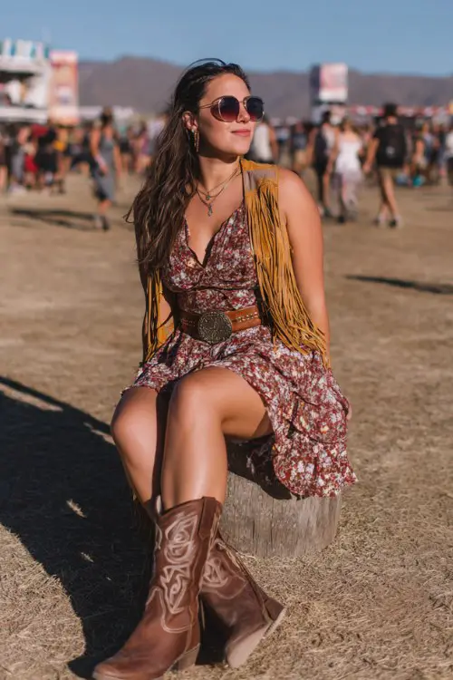 A curvy woman wears brown cowboy boots with a tiered floral mini dress and fringe suede vest, accessorized with a wide western belt and oversized sunglasses