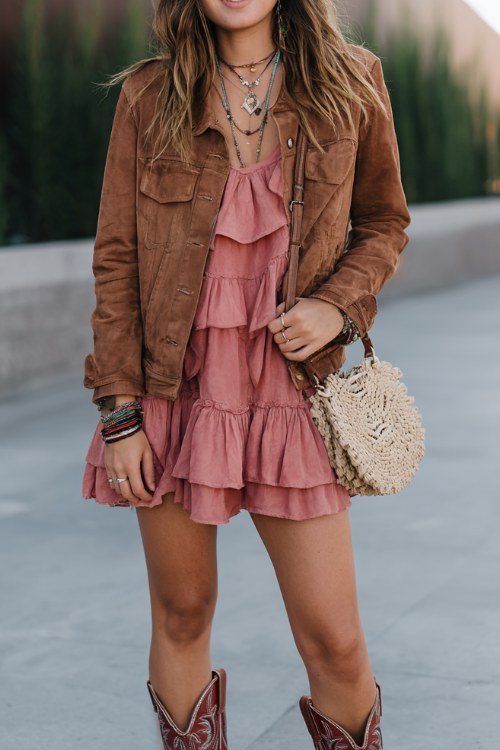 A curvy woman wears dusty pink cowboy boots with a ruffled boho mini dress layered under a suede jacket, accessorized with stacked bracelets