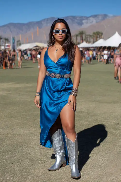 A curvy woman wears silver cowboy boots with a cobalt blue satin slip dress featuring a high slit and western belt