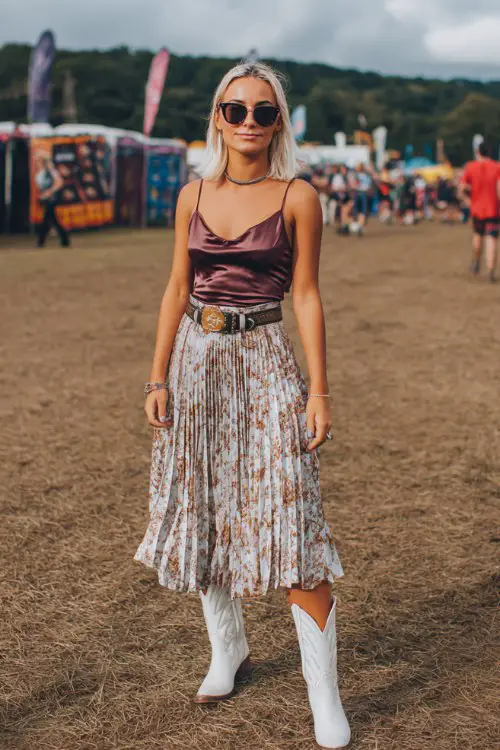 A curvy woman wears white cowboy boots with a pleated midi skirt and fitted satin camisole, styled with a structured belt