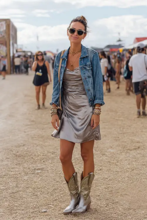 A full body white woman wears metallic bronze cowboy boots with a satin slip skirt and fitted knit top, layered with a cropped denim jacket and bold cuff bracelets