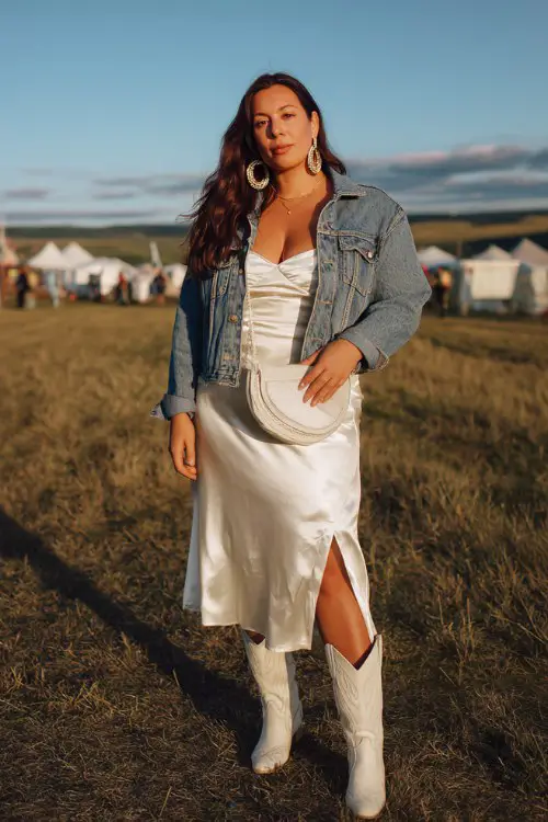 A plus-size white woman wears white cowboy boots with a long satin slip dress layered under a cropped denim jacket, finished with hoop earrings and a sleek crossbody bag