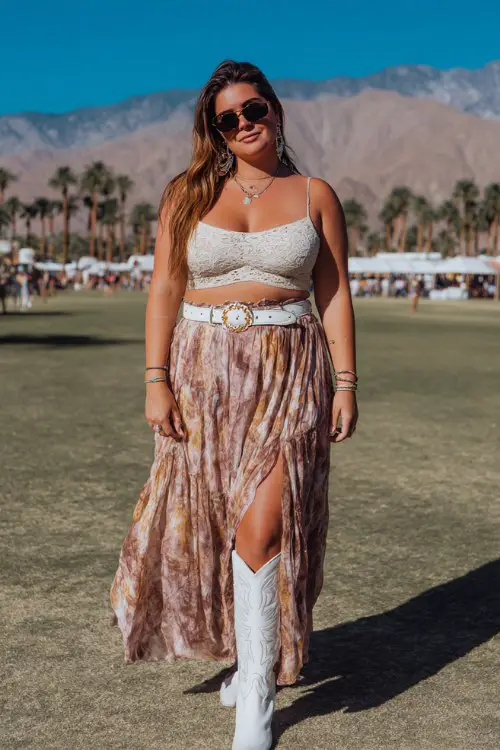 A plus-size woman wears a flowy boho maxi skirt with white cowboy boots, a lace crop top and wide western belt, styled with oversized sunglasses and statement earrings