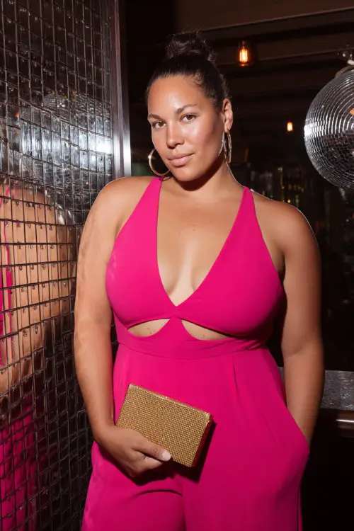 A plus-size woman wears a hot pink cut-out jumpsuit with a slick bun and bold clutch