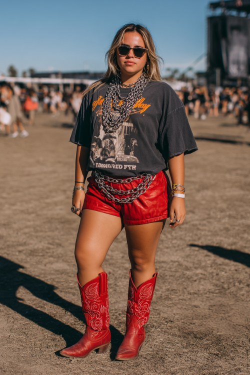 A plus-size woman wears bright red cowboy boots with faux-leather shorts and an oversized graphic tee tucked in, accessorized with layered chains and bold sunglasses