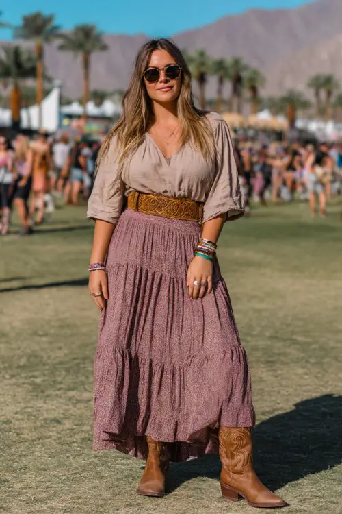A plus-size woman wears brown cowboy boots with a tiered bohemian maxi skirt and a relaxed linen blouse tucked in, accessorized with a woven belt and stacked rings