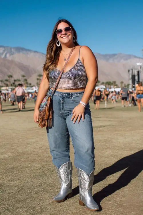 A plus-size woman wears silver cowboy boots with high-waisted flare jeans and a rhinestone mesh crop top layered over a bralette