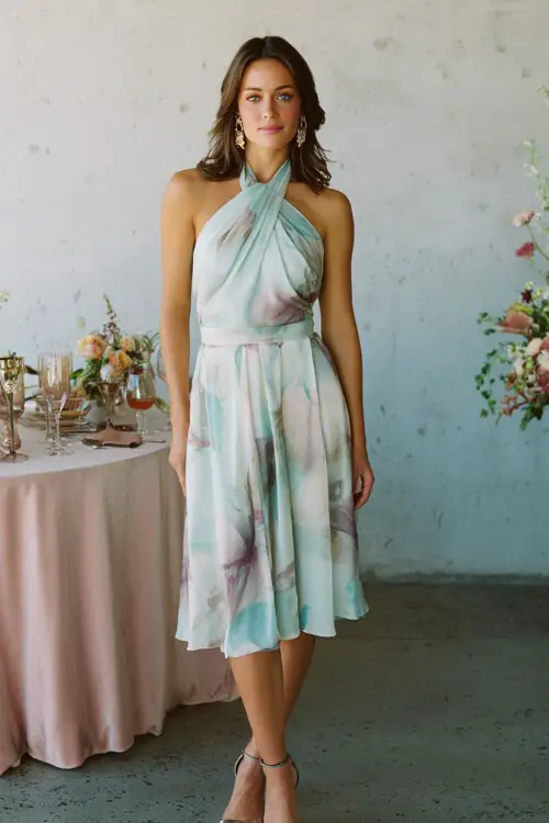 A woman over 30 wears a classy halter-neck cocktail dress with pastel botanical print and elegant pleated skirt, styled with metallic strappy heels