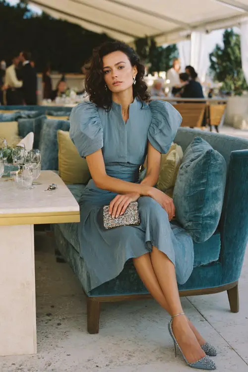 A woman over 30 wears a steel blue midi dress with pleated organza sleeves and pearl buttons down the back, styled with matching heels