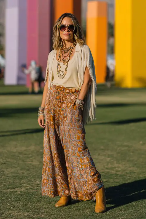 A woman over 30 wears caramel cowboy boots with wide-leg patterned pants and a flowing tunic blouse, accessorized with layered necklaces and oversized sunglasses