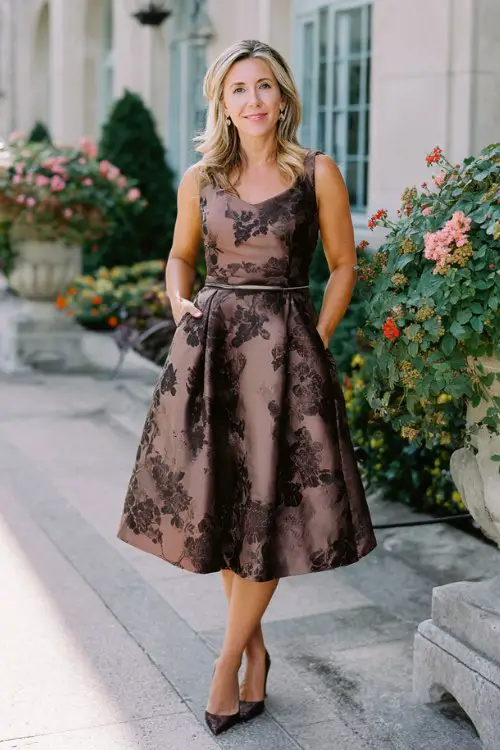 A woman over 40 wears an elegant cocktail dress featuring jacquard floral patterns with subtle metallic threads, tea-length silhouette and delicate belt detail