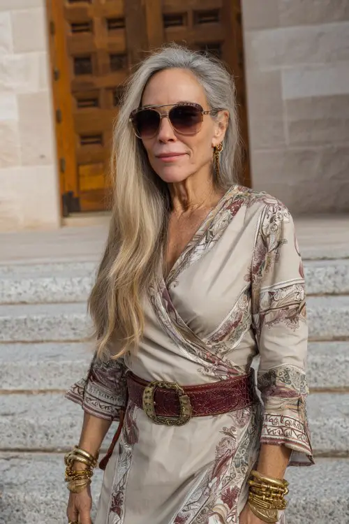 A woman over 40 wears beige cowboy boots with a printed wrap dress and wide western belt, styled with elegant bangles and oversized sunglasses